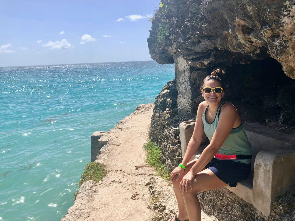 The author sits on a stone bench in a small cave overlooking the ocean in Barbados. She is smiling and wearing yellow sunglasses and a green tank top