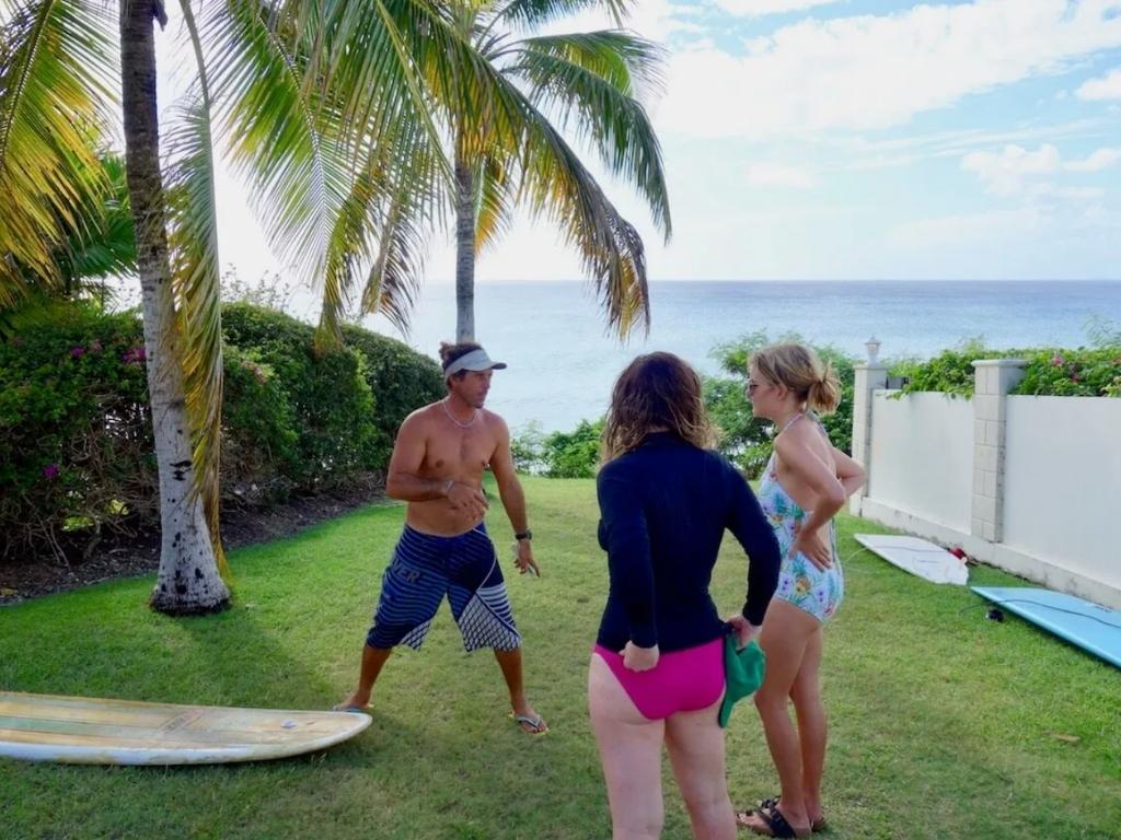 A surf instructor explaining surf technique to two students on a grassy lookout point with the ocean in the background. There are surf boards in the shade of the palm tree at one of the best Barbados surf schools.