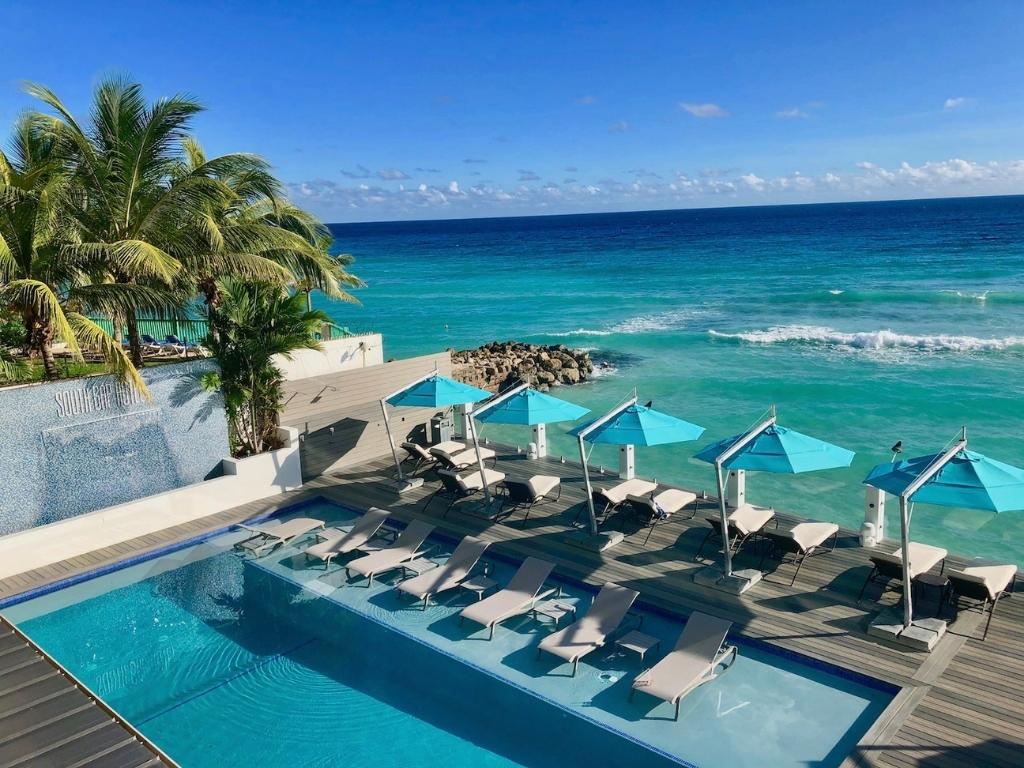 The pool deck at South Gap Hotel in Barbados. The patio extends over the ocean and 15 pool loungers are arranged facing the ocean and the pool.
