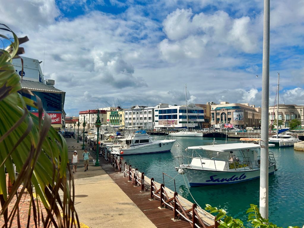 The Bridgetown harbor viewed from the bridge that runs through town. There are boats floating in the foreground and colorful buildings on the other side of the water