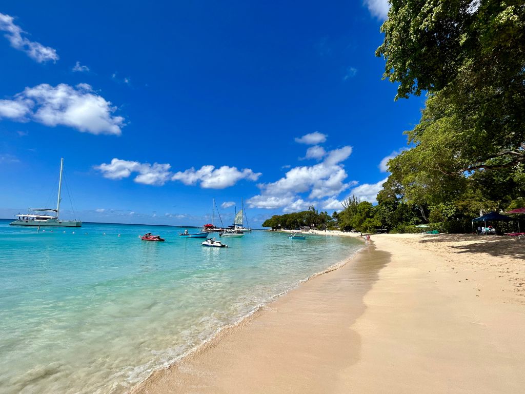 A beautiful beach near Holetown Barbados with white sand and clear blue water. A few boats and jetskis float on the calm waters and trees provide some shade on the beach