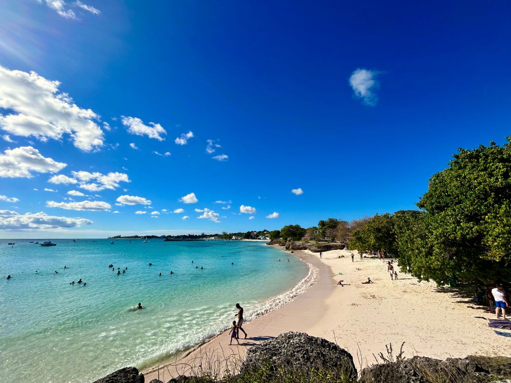 Miami Enterprise Beach in Oistins. A wide white-sand beach with trees providing shade. The water is crystal clear and families play in the gentle waves. If you aren't sure where to stay in Barbados, Oistins might be a good fit