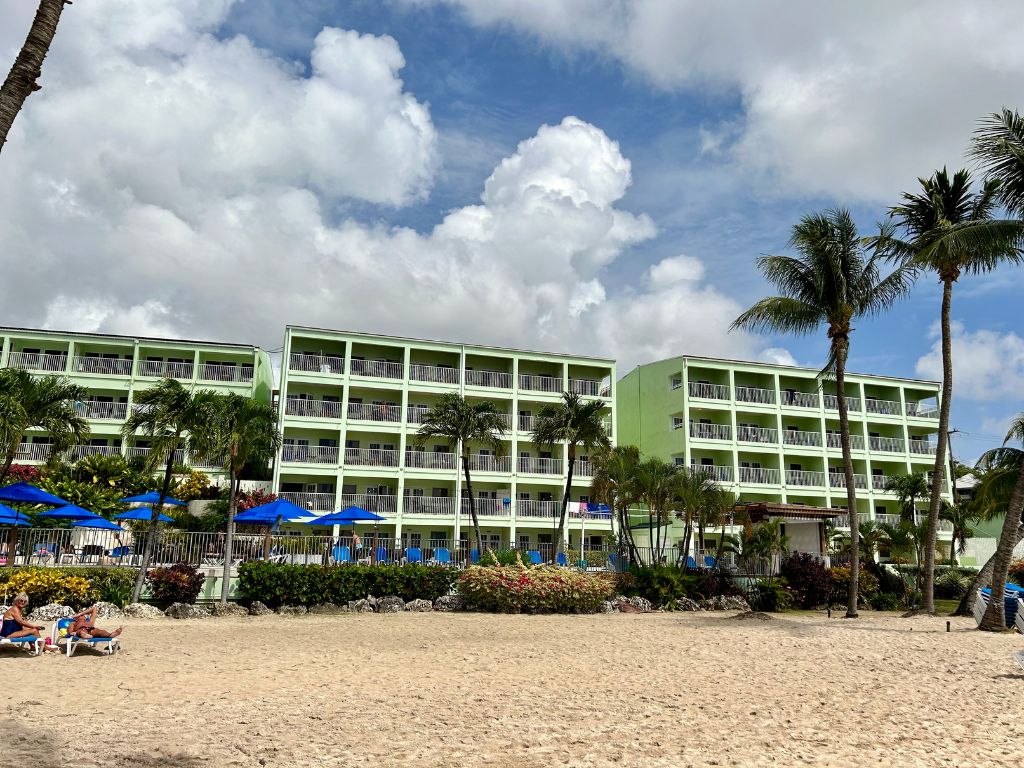 Coconut Court Hotel in Barbados has a green exterior and blue umbrellas in front. There are coconut trees surrounding it and it's a sunny day.